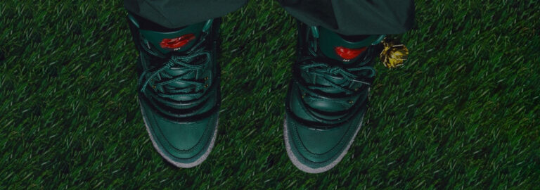 Top-down view of the Air Jordan 3 ‘Concrete Rose’ sneakers on green grass. The shoes feature a deep green leather upper with a vibrant red rose emblem on the tongue and detailing that wraps around the lower shoe to mimic thorns.